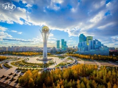 La Torre Bayterek si erge al centro di una piazza circolare circondata da edifici, alberi e auto parcheggiate, sotto un cielo parzialmente nuvoloso a Nur-Sultan. Scoprite questo monumento iconico durante il vostro tour del Kazakistan.