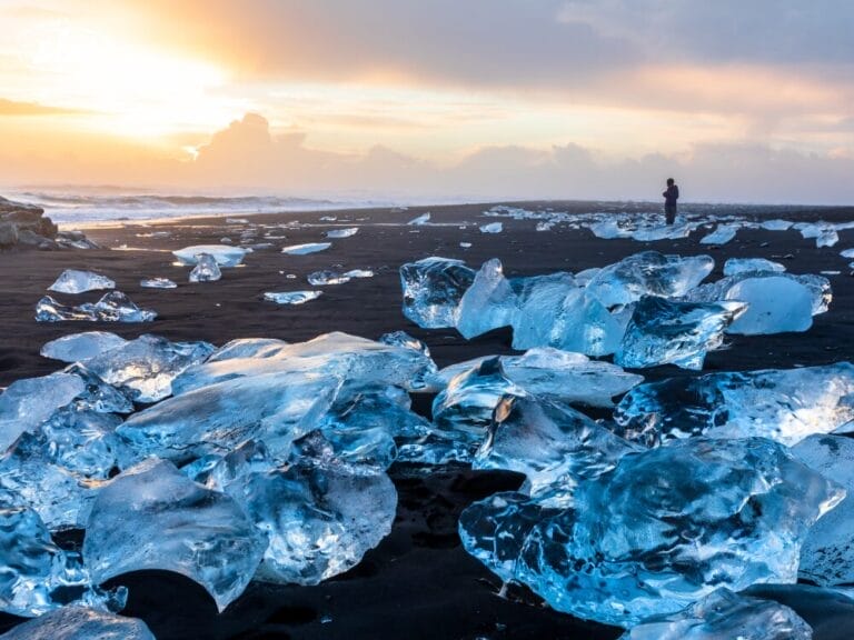 Grossi pezzi di ghiaccio sparsi su una spiaggia di sabbia nera all'alba nel Sud Islanda, con una persona in piedi in lontananza vicino alla riva.