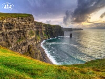Ripide scogliere con pareti rocciose stratificate si affacciano sull'oceano sotto un cielo nuvoloso al tramonto, con le onde che si infrangono in basso e l'erba che ricopre la cima delle scogliere: uno scenario iconico di un'esperienza esclusiva del Tour Irlanda.