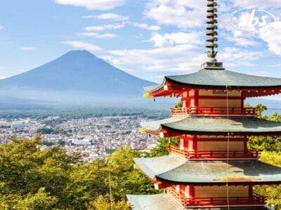 Una pagoda rossa si erge in primo piano, con il Monte Fuji e un paesaggio urbano visibile sullo sfondo sotto un cielo parzialmente nuvoloso, catturando la serena bellezza del Giappone durante la stagione dei Matsuri.