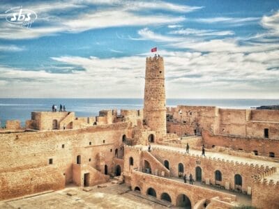 Antica fortezza in pietra con una torre centrale, persone che camminano e il mare sullo sfondo sotto un cielo azzurro: uno spettacolo iconico di ogni avventura del Tour Autentica Tunisia.