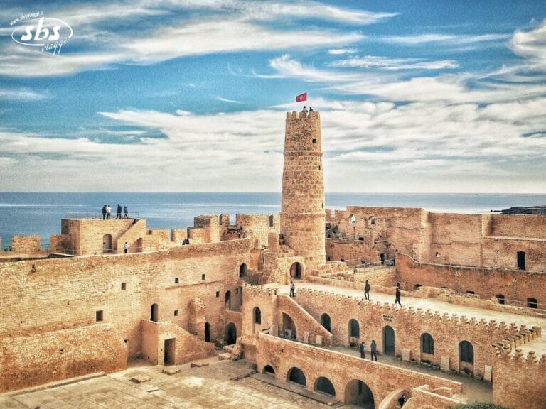 Antica fortezza in pietra con una torre centrale, persone che camminano e il mare sullo sfondo sotto un cielo azzurro: uno spettacolo iconico di ogni avventura del Tour Autentica Tunisia.