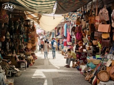 La gente cammina in un vivace mercato all'aperto, molto simile a quelli del Gran Tour del Marocco, fiancheggiato da borse colorate, tessuti e oggetti artigianali su entrambi i lati.