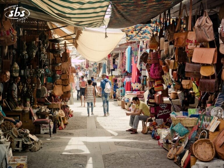 La gente cammina in un vivace mercato all'aperto, molto simile a quelli del Gran Tour del Marocco, fiancheggiato da borse colorate, tessuti e oggetti artigianali su entrambi i lati.