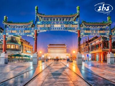 Un arco tradizionale cinese illuminato si erge all'ingresso di una strada, con edifici storici e una grande porta sullo sfondo al tramonto, catturando il fascino del Guizhou descritto da Cina d'Autore.
