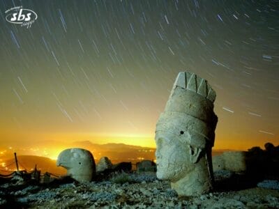 Antiche statue di pietra sul Monte Nemrut sotto un cielo notturno con scie di stelle e luci della città in lontananza: uno scenario magico tratto da Colori della Turchia, perfetto per il tuo prossimo Tour Turchia.