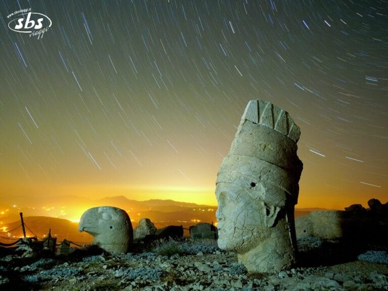 Antiche statue di pietra sul Monte Nemrut sotto un cielo notturno con scie di stelle e luci della città in lontananza: uno scenario magico tratto da Colori della Turchia, perfetto per il tuo prossimo Tour Turchia.
