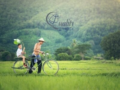 Un uomo e un ragazzo percorrono in bicicletta un campo verde con colline sullo sfondo; il logo "Quality Group" sovrapposto cattura lo spirito di un'esperienza Tour Cambogia.