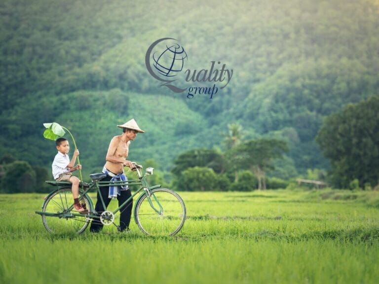 Un uomo e un ragazzo percorrono in bicicletta un campo verde con colline sullo sfondo; il logo "Quality Group" sovrapposto cattura lo spirito di un'esperienza Tour Cambogia.