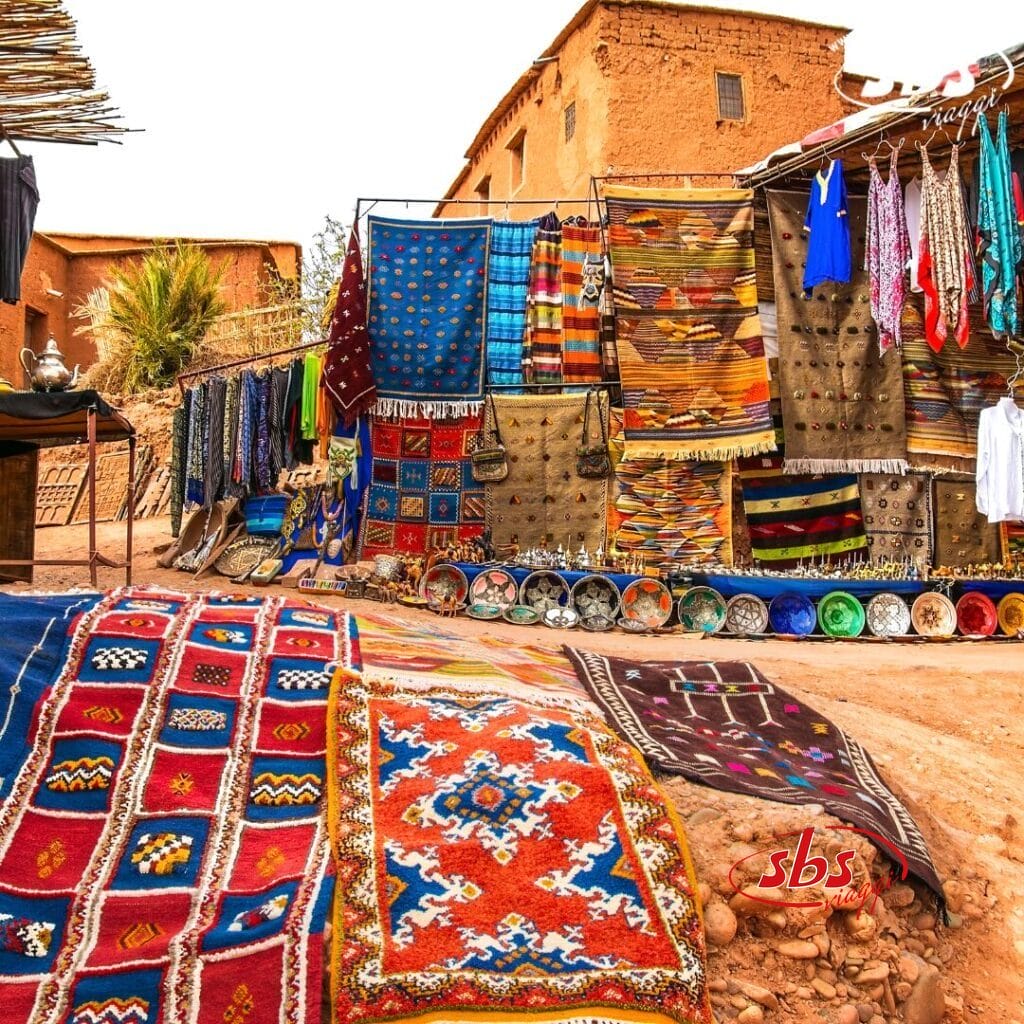 Tappeti intrecciati e ceramiche colorate, parte di una vivace collezione di Bozza automatica, sono esposti fuori da una bancarella di fronte a edifici in adobe.