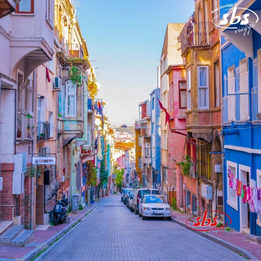 Una strada colorata e stretta, fiancheggiata da auto parcheggiate e vecchi edifici su entrambi i lati, in una città vibrante, cattura l'essenza vivace di Bozza automatica.