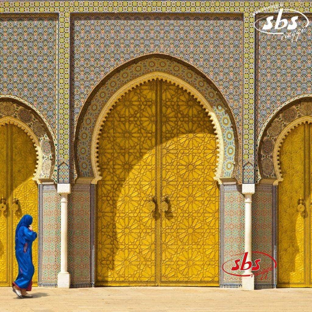 Una persona in vestaglia blu passa davanti a porte dorate ornate con intricati motivi a mosaico, mentre la scena tranquilla evoca un senso di bozza automatica.