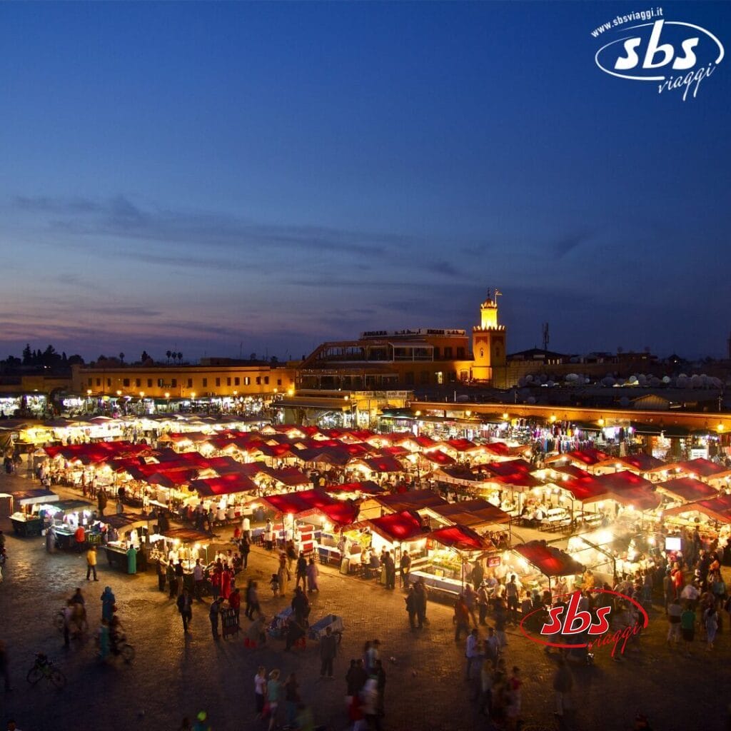 Vista serale di un vivace mercato all'aperto con Bozza automatica, tettoie rosse, luci scintillanti e una torre dell'orologio sullo sfondo.